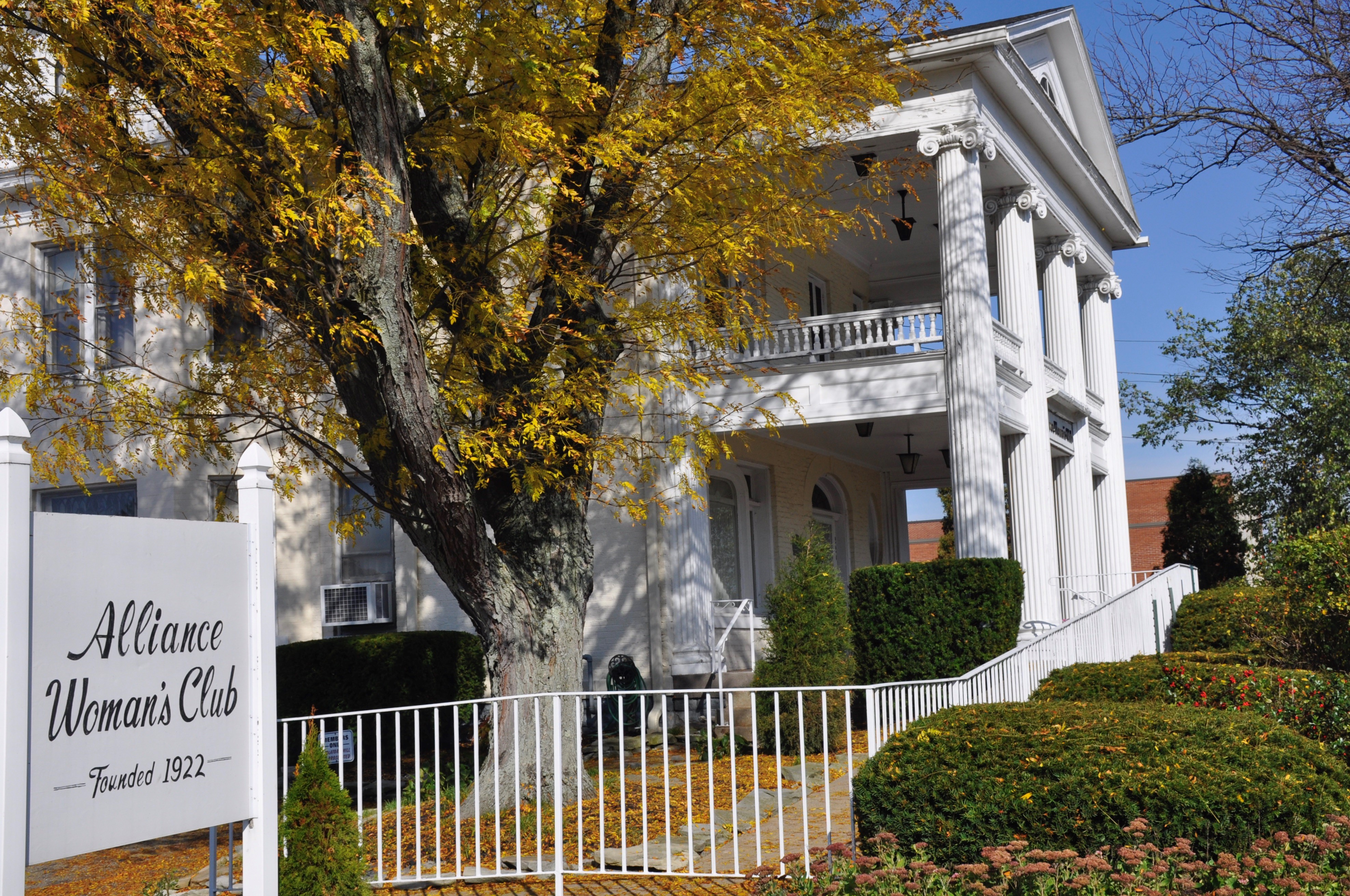 White colonial building with pillars of the Alliance Woman's Club, founded in 1922, amidst autumn foliage.