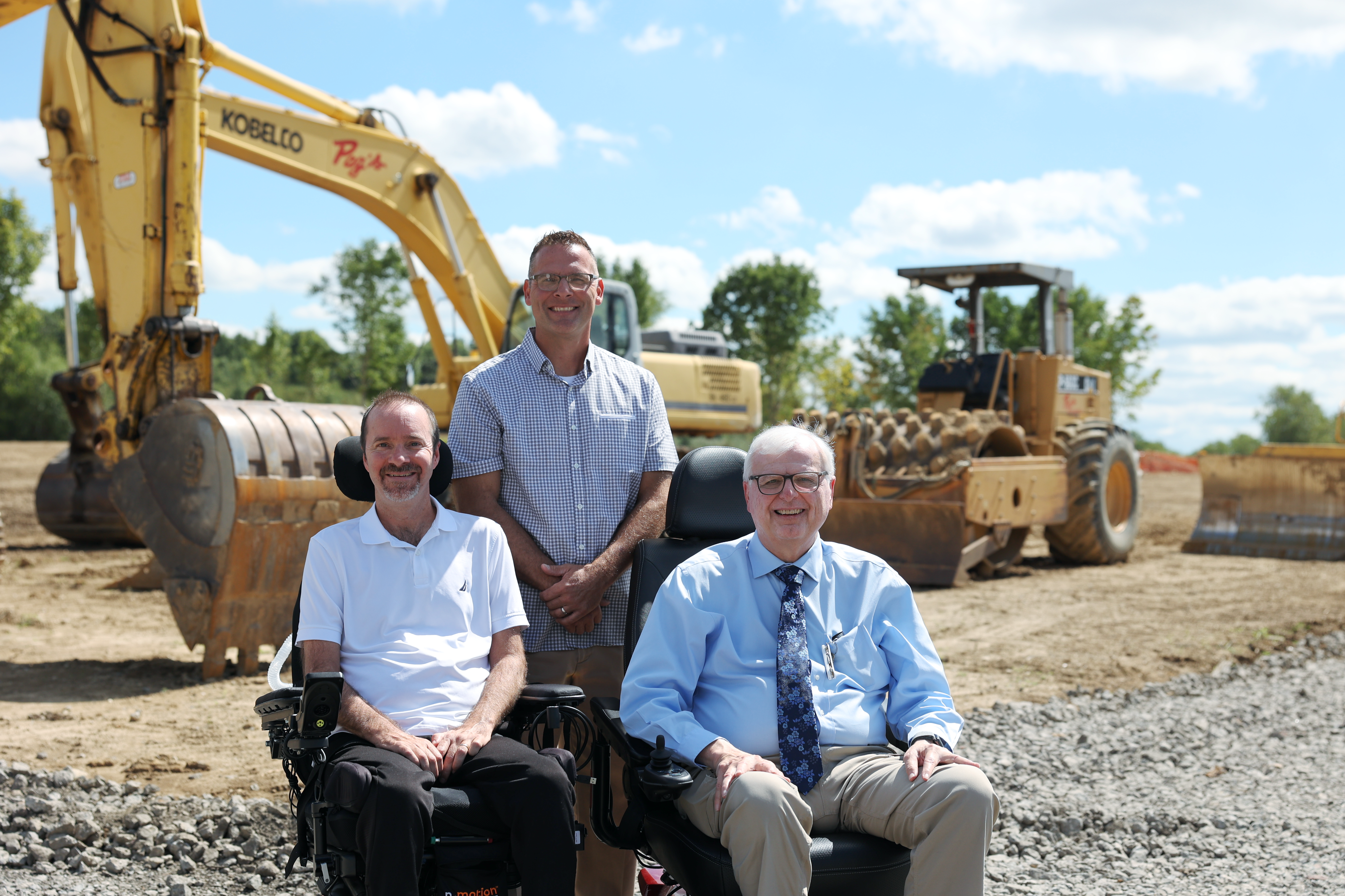 Three smiling men at a construction site, with excavator and bulldozer in the background.